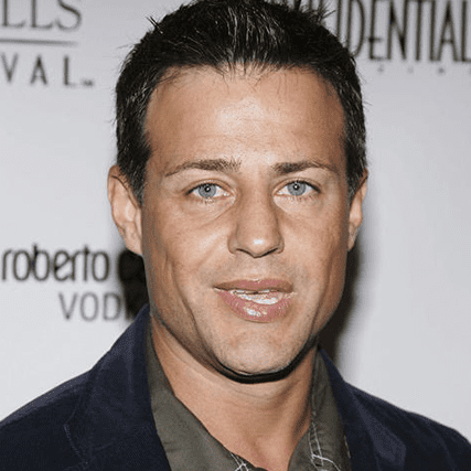 A dark-haired man with blue eyes looks directly at the camera with a subtle smile, wearing a dark jacket against a blurry event background.